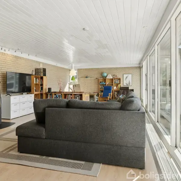 En mørkegrå sofa står foran store vinduer i en stue. En brændeovn står ved væggen, mens en reol med bøger og et skrivebord ses i baggrunden.