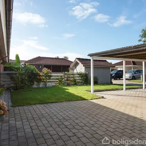 En flisebelagt indkørsel fører til en carport ved siden af et murstenshus med en lille have. Træer og huse findes i baggrunden under en blå himmel.