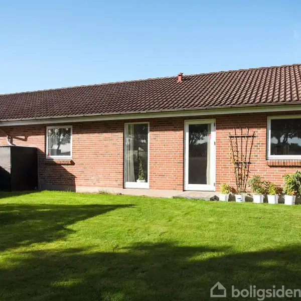 Rødstenshus med skråtag står stille i solen. Foran huset er en velplejet græsplæne med planter og små buske langs ydervæggen. En lille have med drivhus ses til venstre.