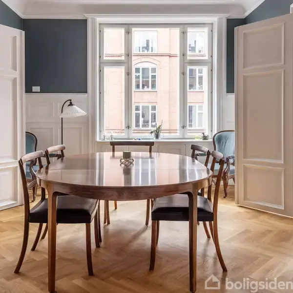 Et rundt spisebord med seks stole står i en stue med sildebensparket. To store vinduer forsyner rummet med lys, og en lampe ses ved siden af bordet.