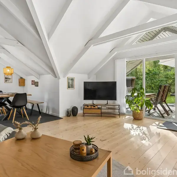 Et moderne opholdsområde med et TV på en lav reol, stueplanter og store glasdøre, der åbner ud til en terrasse. Spisebord med sorte stole i baggrunden, hvide lofter med synlige bjælker.