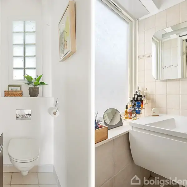 Toilet er placeret i et lyst rum, omgivet af fliser og hylder med dekorationer og en plante. Håndvask og spejl ved siden af et stort matteret vindue, med toiletartikler organiseret ved vasken.