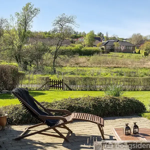 En liggestol står på en terrasse med udsigt over en have og et åbent grønt landskab. I baggrunden ses træer, buske og et hus. To lanterner står ved siden af stolen.