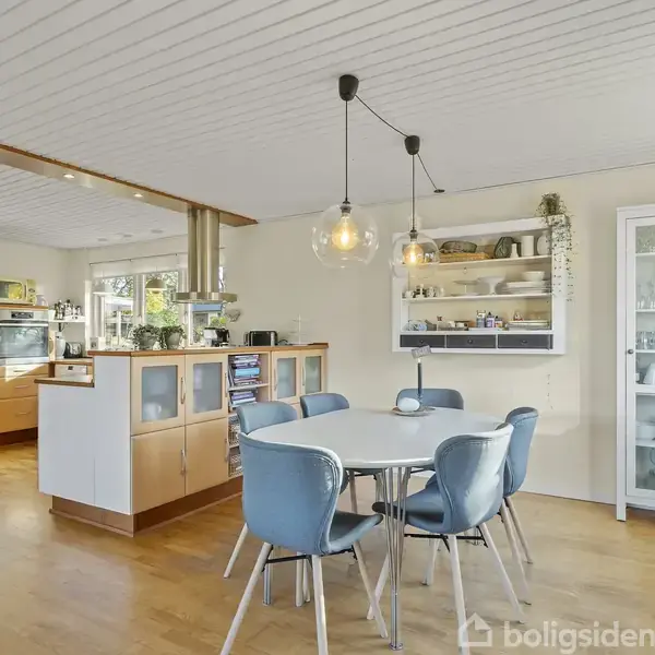 Et moderne køkken- og spisestueområde. Fem blå stole står omkring et rundt spisebord. Hylder med pynt genstande og en glas skab findes i baggrunden. Store vinduer giver naturligt lys.