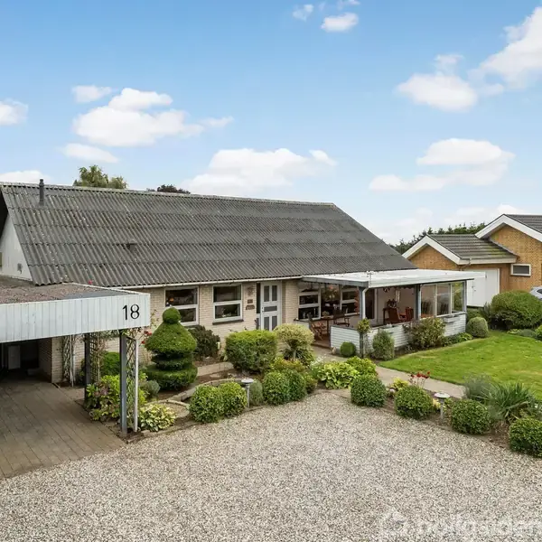 En bungalow med sort tag vises, beliggende ved en grusbelagt indkørsel og omgivet af en velplejet have. Nummer 18 er synligt ved indkørslen. Huset har naboer og er under en blå himmel.