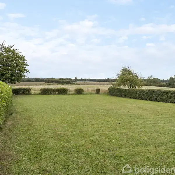 Græsplæne omgivet af klippede hække og enkelte træer, strækker sig mod marker under en let skyet himmel.