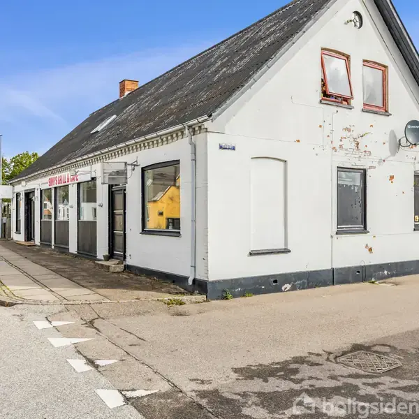 En hvid, ældre bygning med skråt, gråt tag står på et gadehjørne. Et skilt lyder "Grill House." Bygningen er omgivet af et fortov og en vej, med træer i baggrunden.