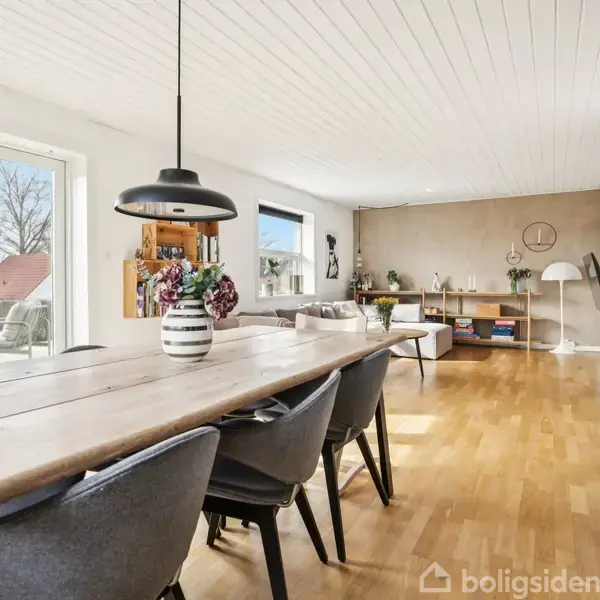 Spisebord med sorte stole står i en lys stue med trægulv. En sort pendel hænger over bordet. Reol og lampe i baggrunden. Store vinduer til venstre.