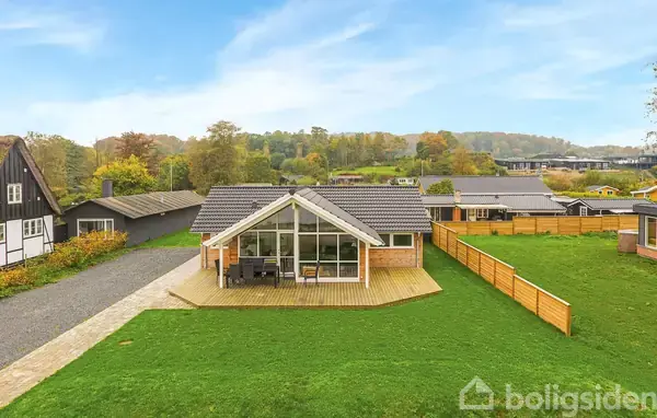 Rødt murstenshus med store glaspartier, står på en grøn græsplæne. Huset har en træterrasse foran og er omgivet af et træhegn. I baggrunden ses flere huse og træer.