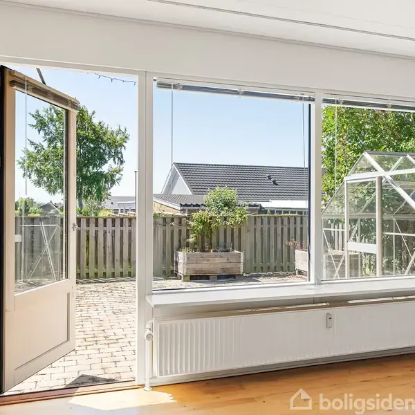 Dør står åben indtil terrasse med flisebelægning og lille drivhus. Indendørs ses et parketgulv og en radiator. En træhæk og blå himmel udgør baggrunden.