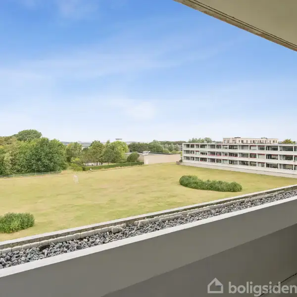 Lejlighedsbalkon med udsigt over en græsplæne og en beboelsesblok i baggrunden. Omgivet af buske og træer under en klar blå himmel.