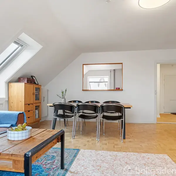 Stue med skråloft, hvor en sofa og sofabord står ved vinduet. Et spisebord med fire stole er placeret bagved. Reoler med bøger står til højre i rummet. Ingen tekst fundet.