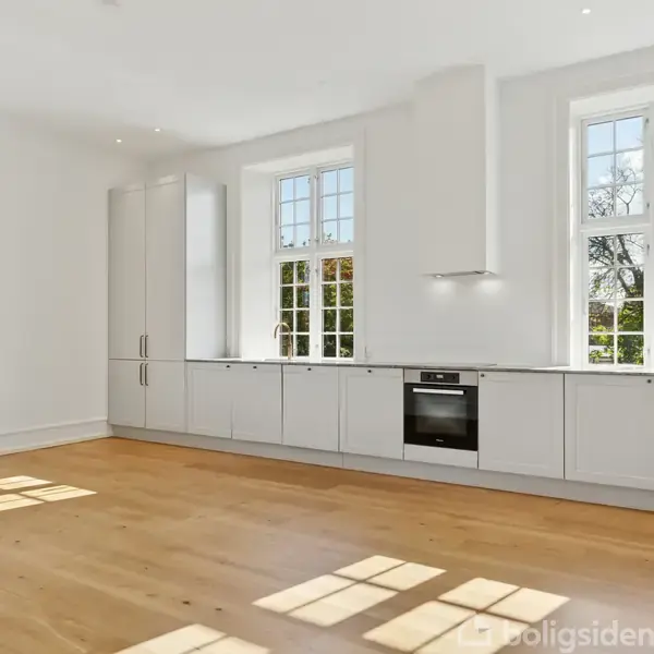 Et moderne køkken med hvide skabe, indbygget ovn og komfur, står opstillet langs en væg. Store vinduer og trægulv skaber et lyst og rummeligt miljø.