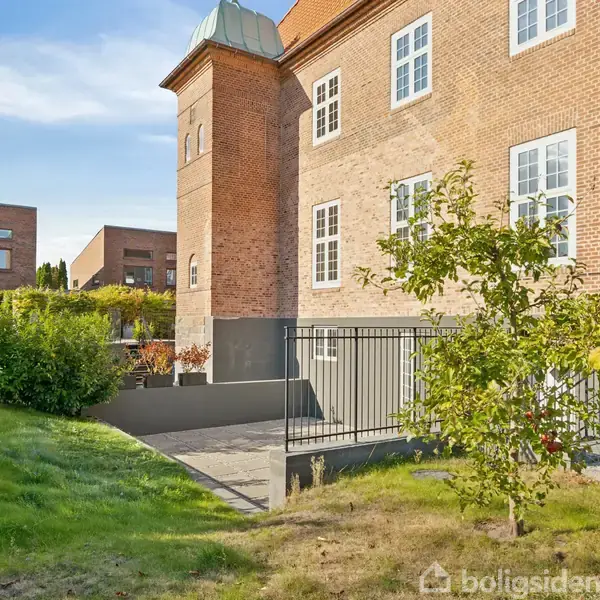 En murstensbygning står stille i et forstadskvarter. En velplejet grøn have omgiver den, med buske og små træer. Sollys belyser facaden og skaber et fredfyldt udseende.