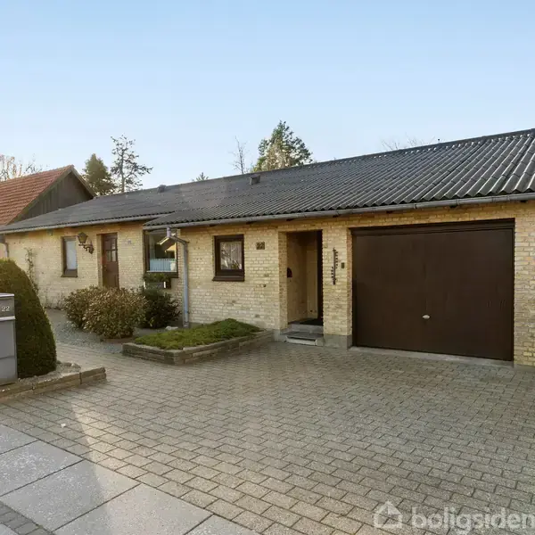 Et murstenshus med garage står på en flisebelagt indkørsel. Huset er omgivet af haveplanter. Et skilt ved vejen til venstre viser teksten "TIL SALG".