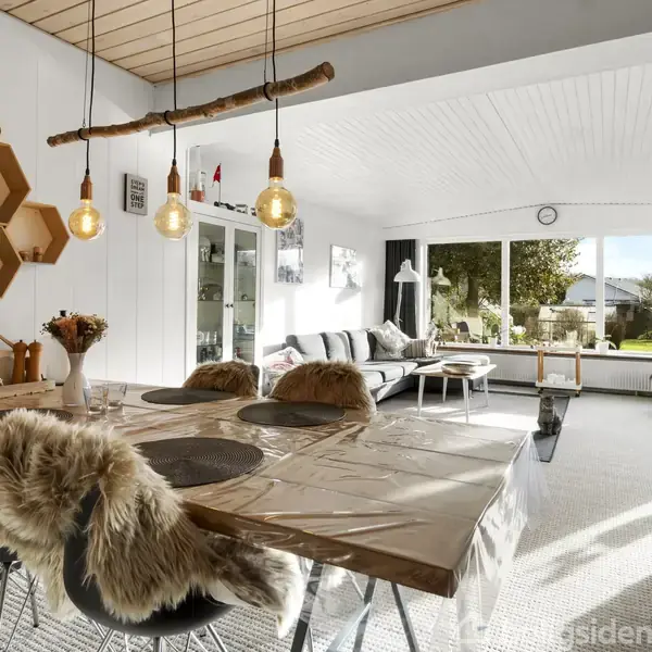 Spisebord dækket med skindbetrukne stole står i moderne stue med lyse lamper hængende fra trægren. Stuen har vinduer mod have, sofaer og dekorative hylder med geometrisk design.