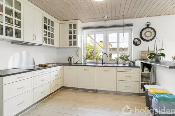 Lyst køkken med hvide skabe i hjørnet, moderne vandhaner og overhængende loftslamper. Stort vindue med udsigt til et hus og have. Dekorativ vægur og planter til højre.