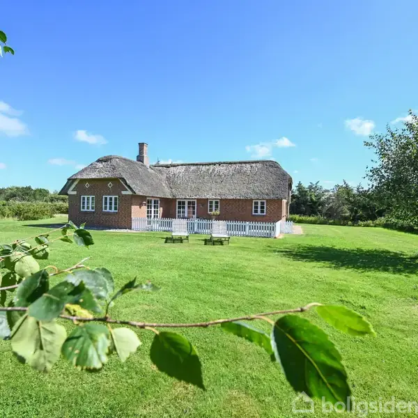 Et stråtækt hus står stille på en stor, grøn græsplæne under en klar blå himmel. Buske og et træ omkranser huset, hvilket skaber en idyllisk landlig atmosfære.