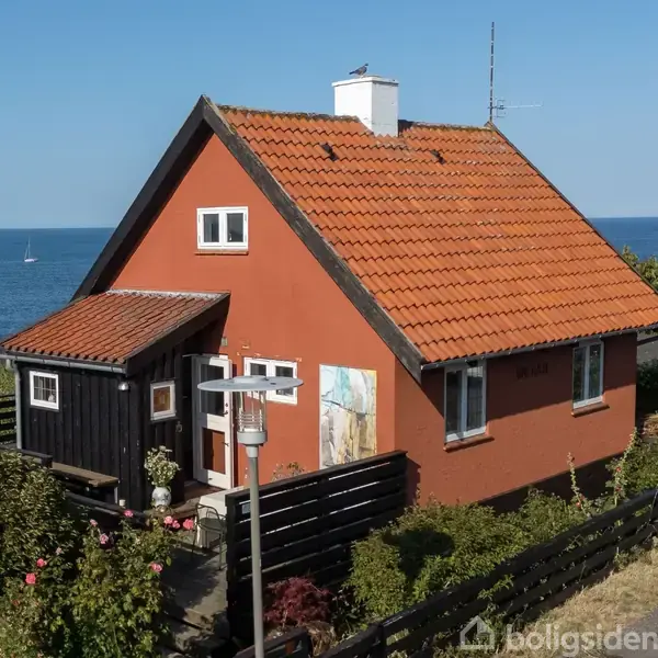 Et rødt hus med skråtag og hvid skorsten står ved havet. En båd sejler i baggrunden. Huset er omgivet af en have og en lille sort havelåge.