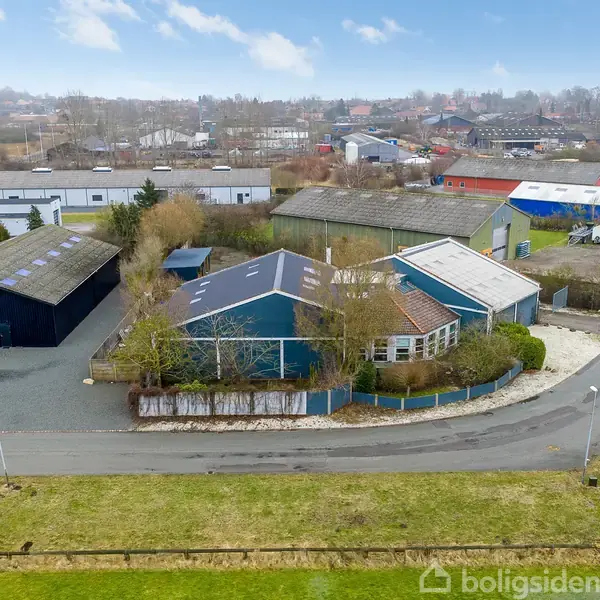 Bygninger i et industriområde med flere lagerbygninger, parkerede biler og buske omkring. I baggrunden ses en by med flere bygninger. Himlen er lys og let skyet.
