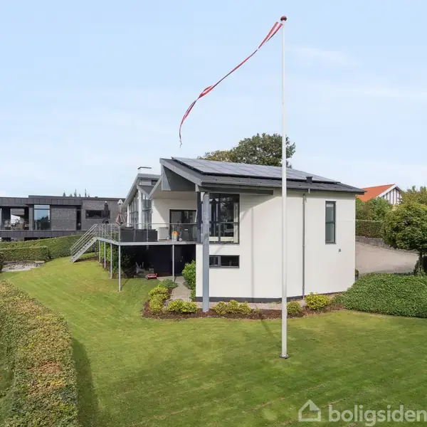 En pejs med brændende ild hænger på en væg i et moderne stueinteriør med store vinduer. Udsigten viser en sø omgivet af huse og frodige grønne områder.