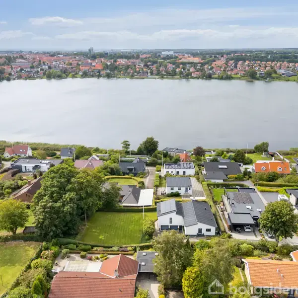 Huse står stille langs en søbred i et beboelseskvarter. Søen er i baggrunden med en by i horisonten, omgivet af træer og græsplæner.