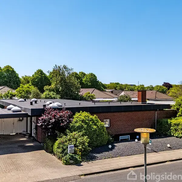 En etplans murstensvilla står i et beboelsesområde med træer og hække. Huset har fladt tag, carport og indkørsel med brosten. Blå himmel danner baggrund.