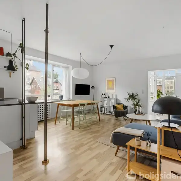 Moderne stue med lyse trægulve, hvor en sofa vender mod et rundt sofabord. I baggrunden ses et spisebord med stole. Rummet er lyst med store vinduer og minimalistisk indretning.