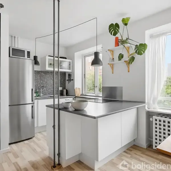 Et moderne køkken med stål køleskab, hvid køkkenø og sort lampe. Overfladen er grå med skål. Vinduer slipper lys ind, og en plante hænger som dekoration.