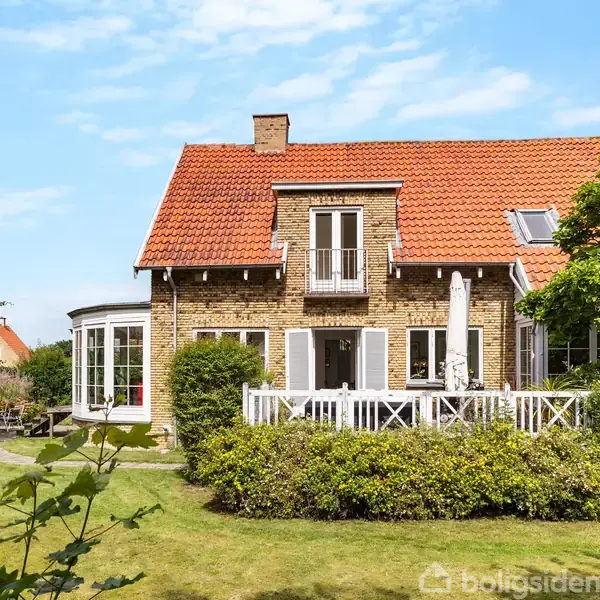 Et murstenshus med rødt tegltag har en veranda med havemøbler. Det står i en velholdt have med grønne buske og træer, under en blå himmel.