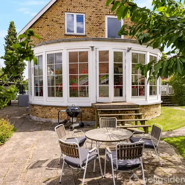 En rund bygning med store vinduer står i en have. En udendørs siddegruppe og grill findes på en fliseterrasse. Blomsterbede og træer omgiver området i solskin.