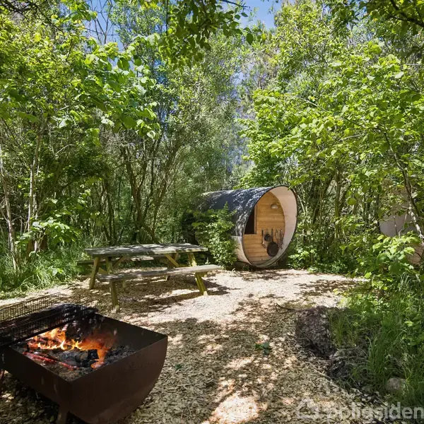 Bålstedet brænder, omgivet af en picnicbænk og en tøndesauna i en frodig, skovklædt have. Sollys siver gennem trækronerne, hvilket skaber en hyggelig atmosfære.