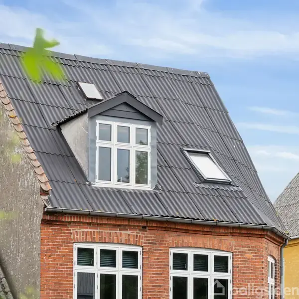 Et rødt murstenshus har en sort skråtag med to vinduer, det ene er et tagvindue. Omgivet af en blå himmel og nabohuse i forskellige farver.