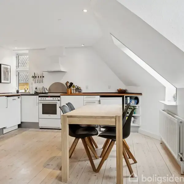 Køkken med hvidt interiør og træbord med stole. Rum indeholder en ovn, opvaskemaskine og køkkenbord ved siden af skråvindue. Hvide vægge og parketgulve skaber en lys atmosfære.