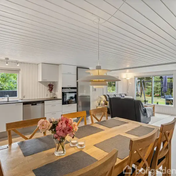 Et lyst køkken-alrum med spisebord fyldt med blomster og lys, åbent ud mod en stue med sofaer og store vinduer, der giver udsigt til en grøn have.