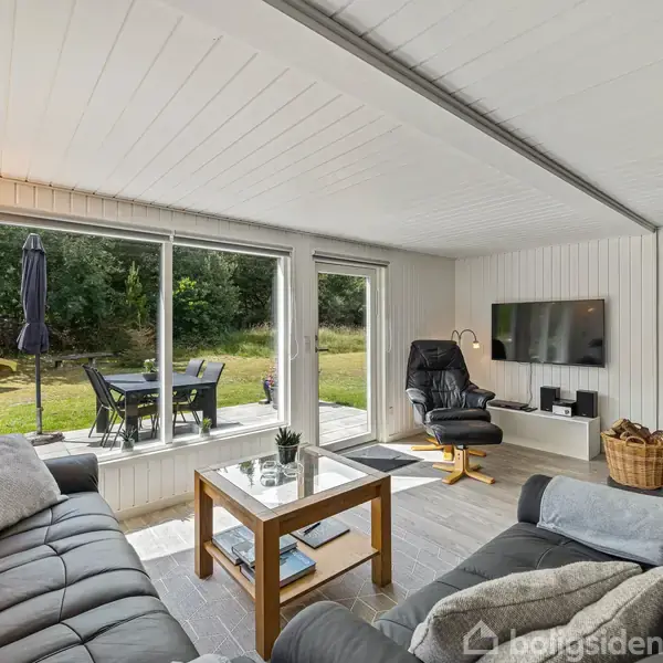 Stue med sorte lædersofaer omkring et sofabord. Et sortbrændt komfur står ved siden af en lænestol, mens store vinduer afslører en terrasse med havemøbler og grøn græsplæne udenfor.