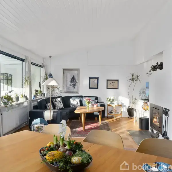 Stue med sort sofa og sofabord, der står foran store vinduer. Til højre en brændeovn, overfor spisestuebord dekoreret med planter og blomster i en lys, indbydende atmosfære.