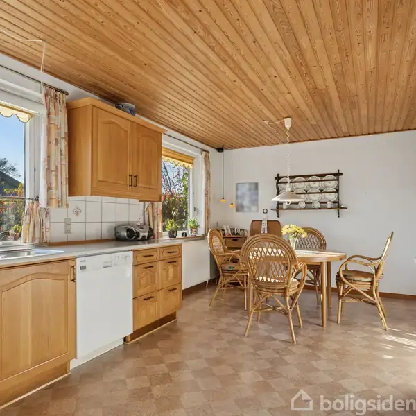 Køkken med træloft og flisegulv, indeholder køkkenbord, vask, opvaskemaskine, og spisebord med fire stol. Store vinduer giver udsigt til en have. Væghængt porcelænsreol og mikroovn ses også.