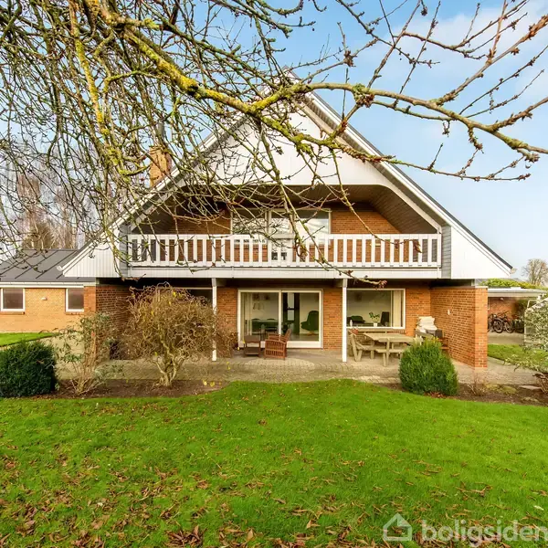 Et rummeligt hus står omgivet af en velplejet have med grønne græsplæner og hække. Huset har en stor balkon og en terrasse. Træer strækker sine grene opad mod en klar himmel.