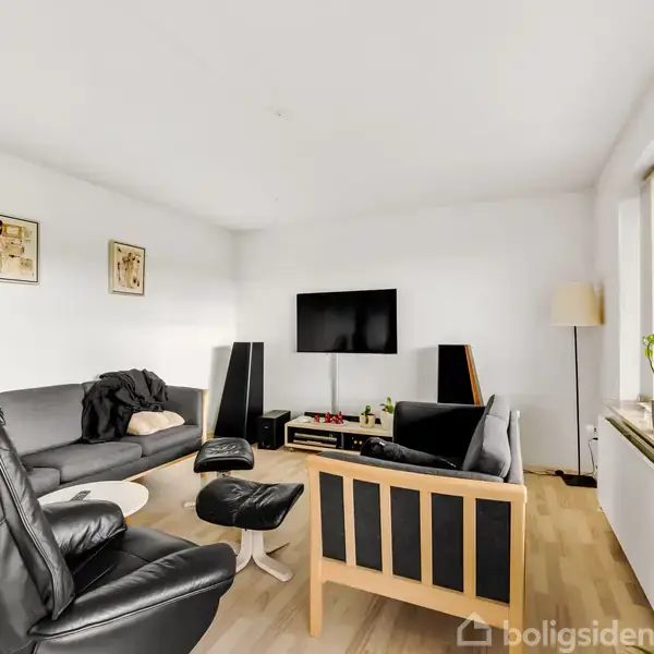 Stue med sorte læderstole og sofa arrangeret omkring et fladskærms-tv på væggen; store vinduer til højre skaber lys, og grønne planter står på vindueskarmen. Tre små kunstværker hænger på væggen.