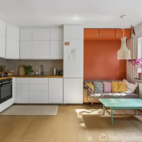 Køkkenalrum med hvidt køkken til venstre, sofa med puder og orange væg til højre. Et vindue med planter bringer lys ind. Grøn kaffebord og moderne lampe supplerer rummet.