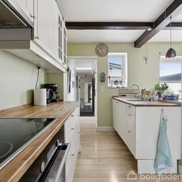 Et lyst køkken med træbordplader og hvide skabe. En kogeplade ses tæt på. Et ur hænger på væggen. Der er masser af lys fra store vinduer.