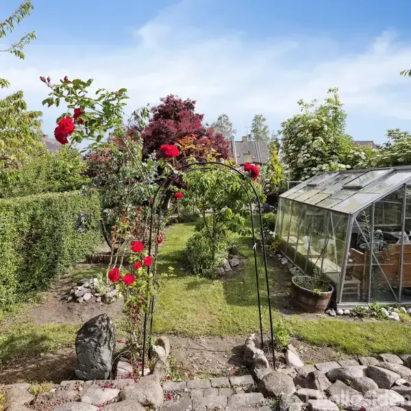 Et drivhus står i en frodig have omgivet af grønne træer og buske. Røde blomster blomstrer på en bueformet støtte. En græsplæne strækker sig i baggrunden under en blå himmel.