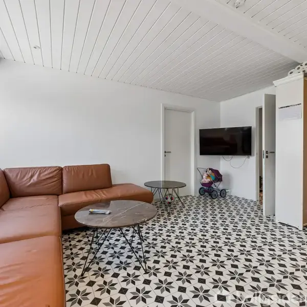 Brun hjørnesofa står i en lys stue med mønstret gulv. To runde sofaborde foran sofaen. Fladskærms-TV hænger på væggen ved legetøjsvogn og hvide skabe.