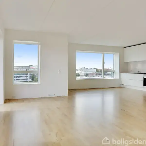 Lyst rum med trægulv, indeholdende et moderne køkken med hvide skabe og indbygget ovn. Store vinduer giver udsigt til bylandskab og slipper naturligt lys ind.