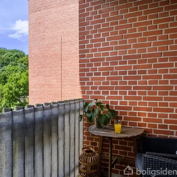 Balkonen har et rundt bord med en plante og et glas juice. To stole er placeret ved siden af. Murstensvægge omgiver balkonen, og i baggrunden ses træer og en bygning.