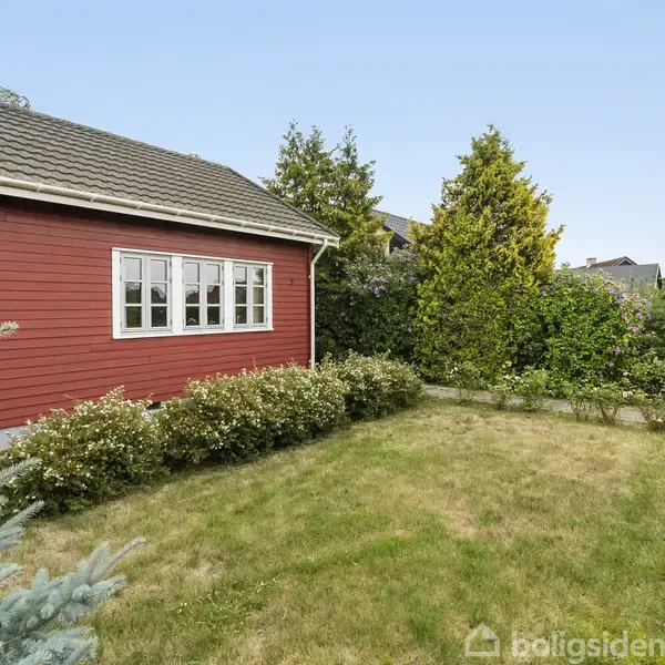 Et rødt hus med hvide vinduer står ved siden af en velholdt have med grønne buske og græsplæne. En sti og hegn ses i baggrunden omgivet af træer.