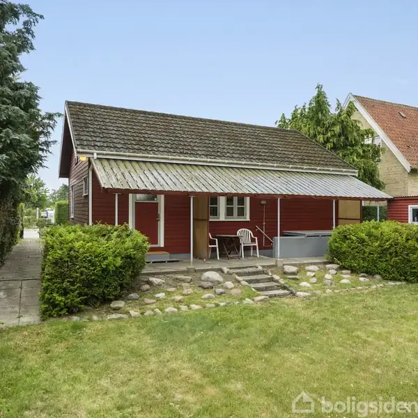 Et rødt træhus med et valmtag står i en grøn have. En lille terrasse har et bord med stole. Stenbelægning og buske omkranser huset.