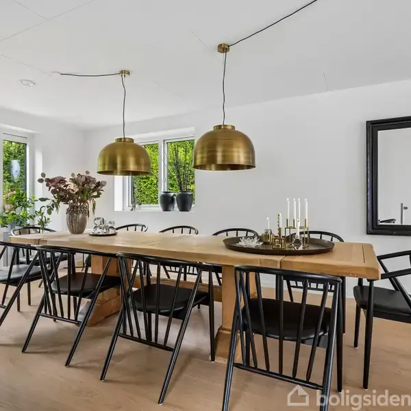 Langt træbord omgivet af sorte stole står i spisestue med hvidmalede vægge. To gyldne loftslamper hænger over bordet. Store glasdøre og et stort vindue giver lysindfald.