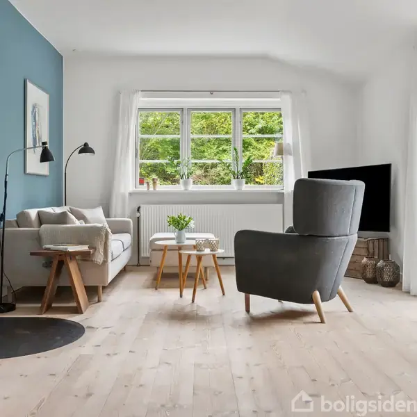 Stue med en grå sofa og lænestol, et væghængt brændeovn der er tændt, stående på et trægulv. Store vinduer giver udsigt til en grøn have.